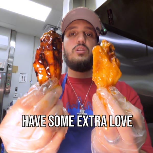 A man in a kitchen holds up two large, saucy chicken wings, one dark and one orange.