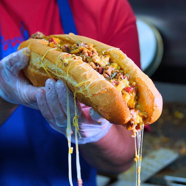 A gloved hand holds a Buffalo Eatz cheesesteak with melted cheese dripping from the sandwich.