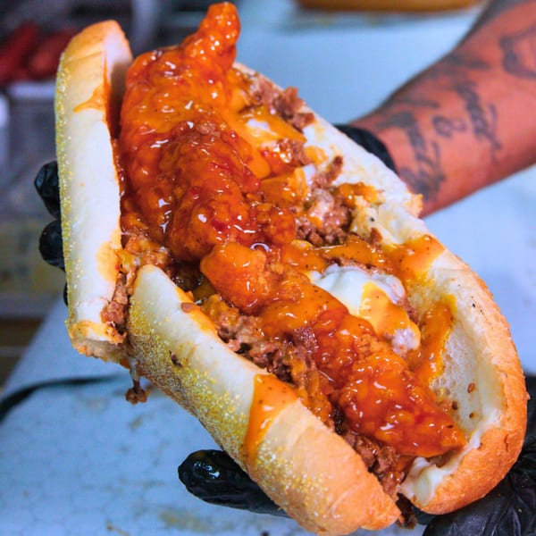 A person in black gloves holds a Buffalo Eatz cheesesteak with chicken tenders and orange sauce.