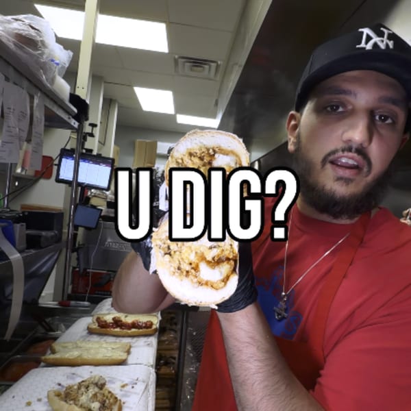 A man in a Buffalo Eatz hat and apron holds up a large chicken sandwich in a kitchen.