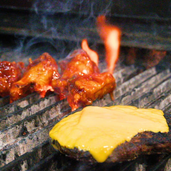 Buffalo Eatz burger patty with melted cheese and buffalo wings cooking on a grill with flames.