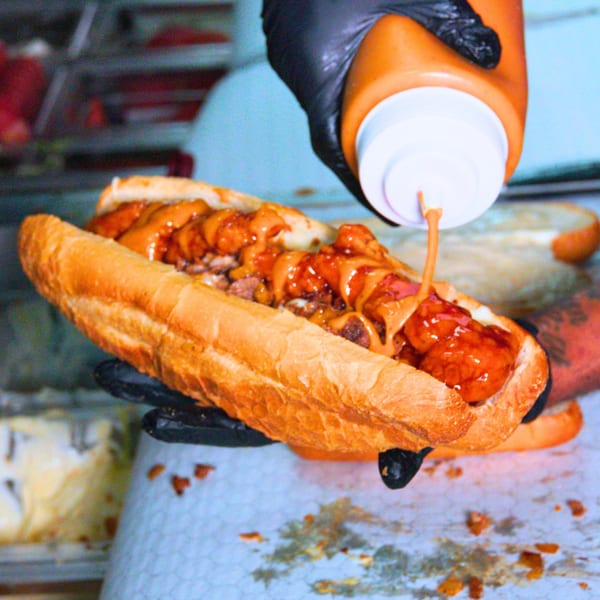A hand in a black glove squeezes orange sauce onto a Buffalo Eatz chicken sandwich.