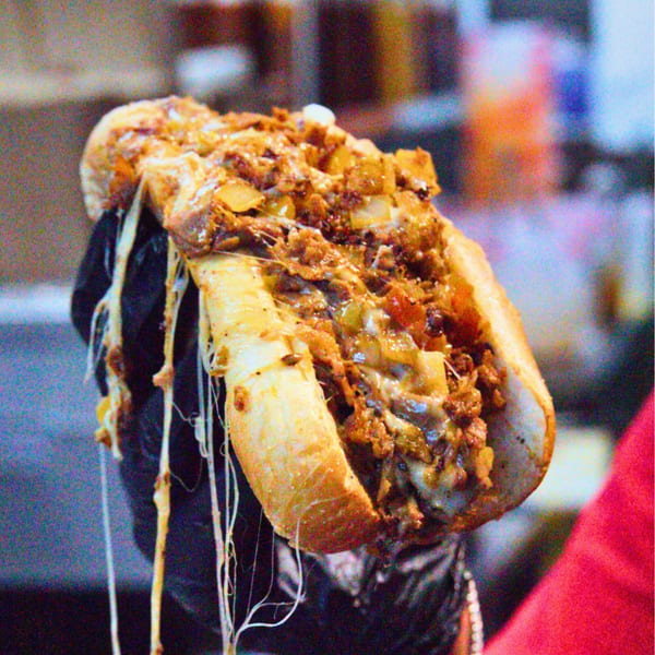 A Buffalo Eatz cheesesteak sandwich, overflowing with meat, cheese, and onions, held by a gloved hand.