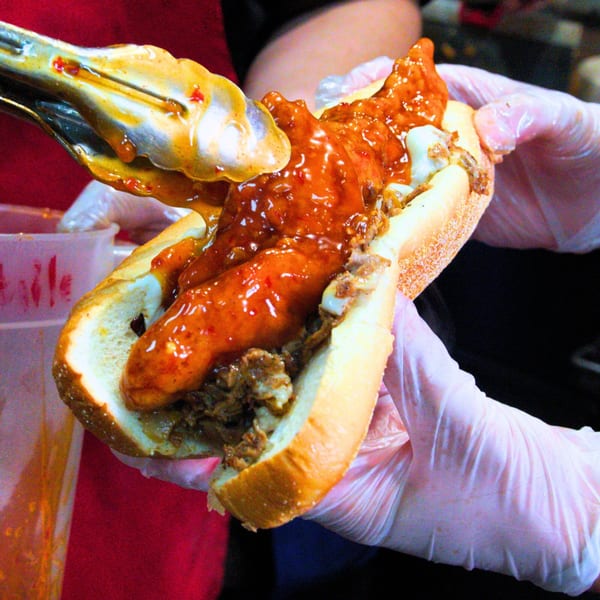 Tongs adding saucy buffalo chicken to a cheesesteak sandwich held by gloved hands.